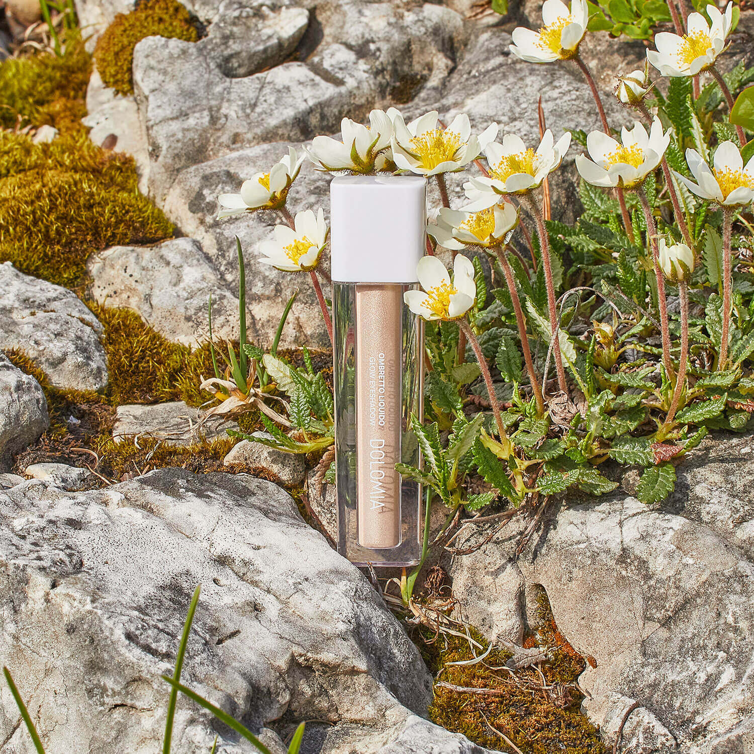 Eine Röhre von Flüssiger Lidschatten (3 Farbvarianten) mit weißer Kappe steht aufrecht zwischen weißen Blüten, grünen Pflanzen und grauen Felsen und hebt ihre langanhaltende Formel und schimmerndes Finish hervor.