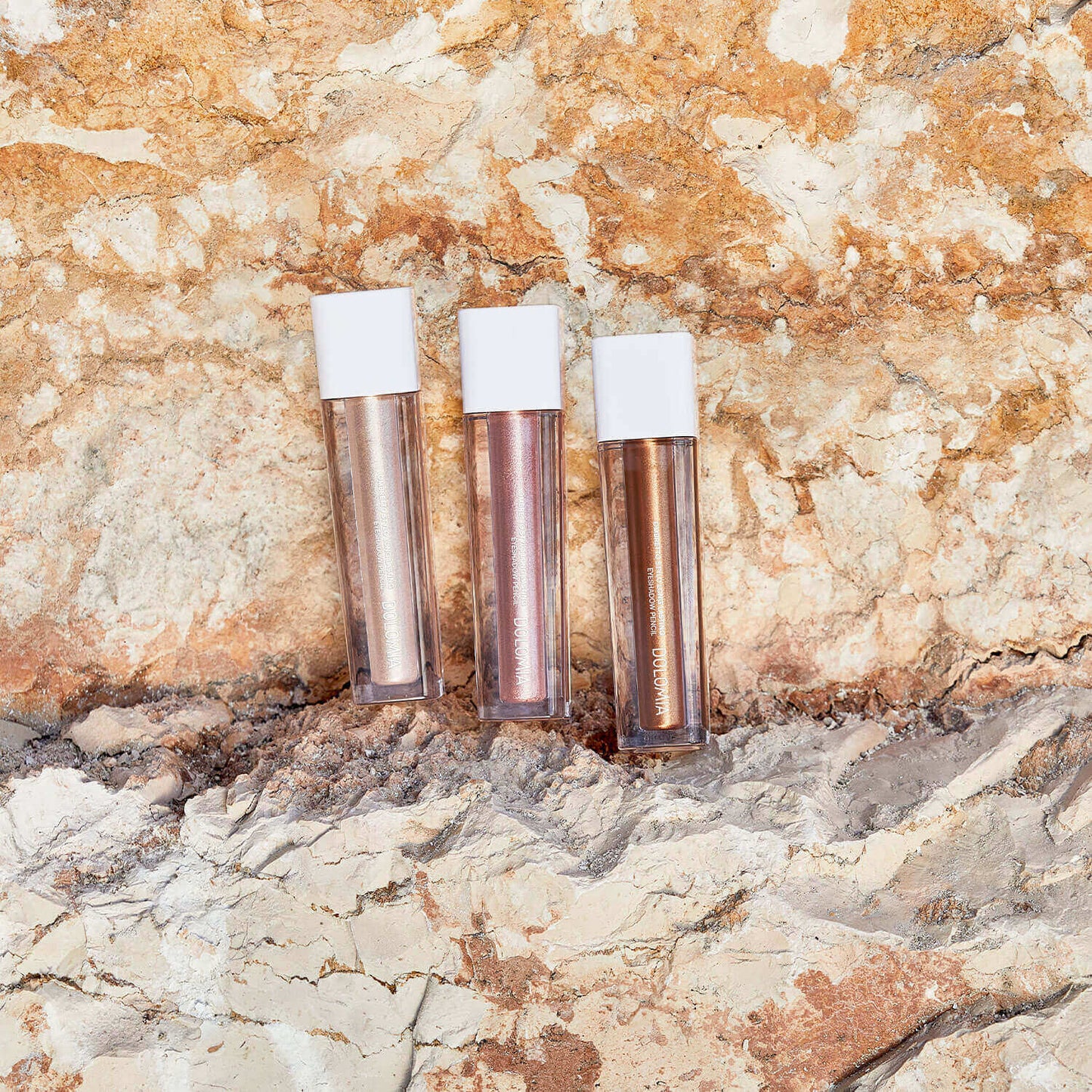 Drei Röhren von Flüssiger Lidschatten (3 Farbvarianten) mit weißen Kappen stehen aufrecht auf einem sandigen, strukturierten Felsen in Erdtönen und bieten jeweils ein schimmerndes Finish.