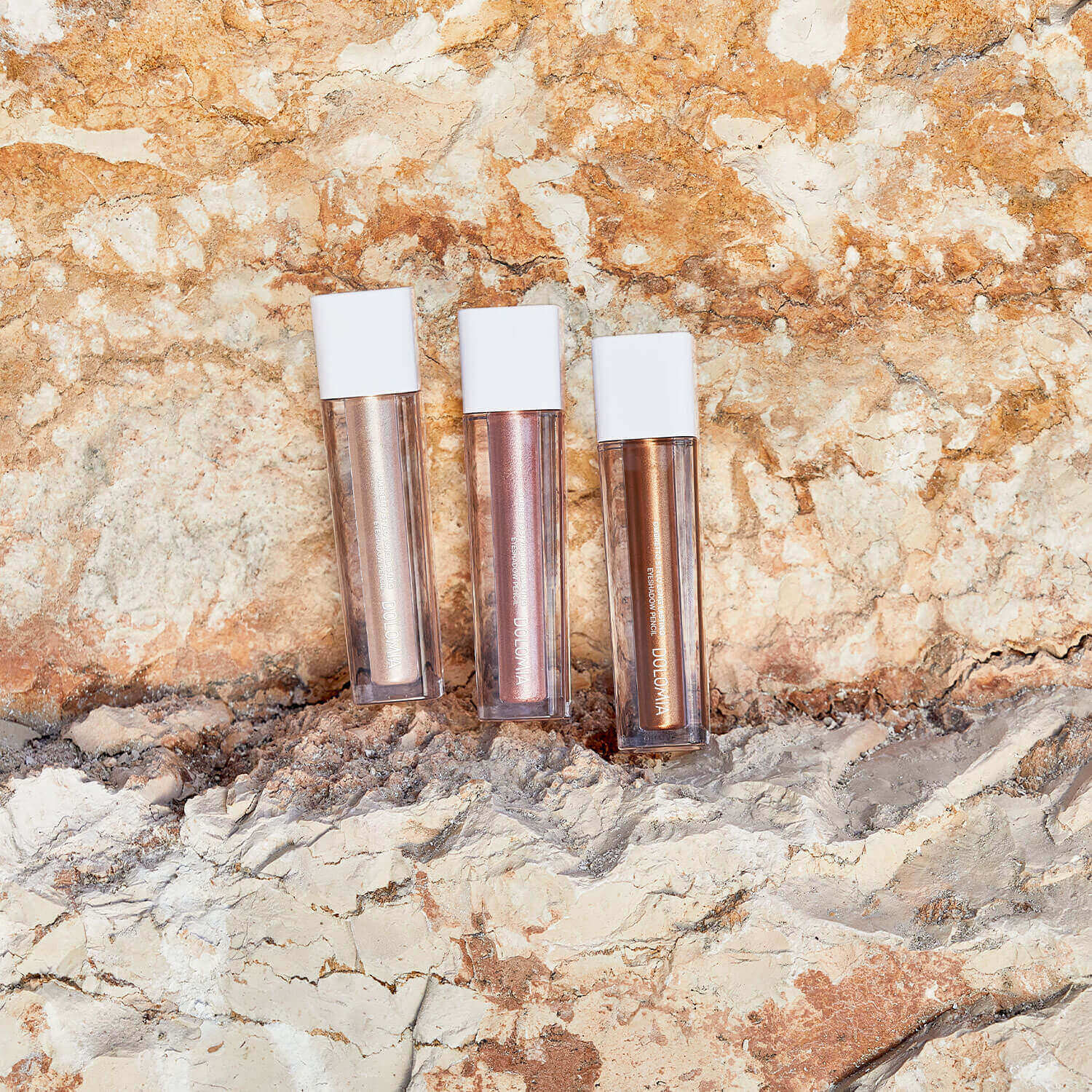 Drei Röhren von Flüssiger Lidschatten (3 Farbvarianten) mit weißen Kappen stehen aufrecht auf einem sandigen, strukturierten Felsen in Erdtönen und bieten jeweils ein schimmerndes Finish.