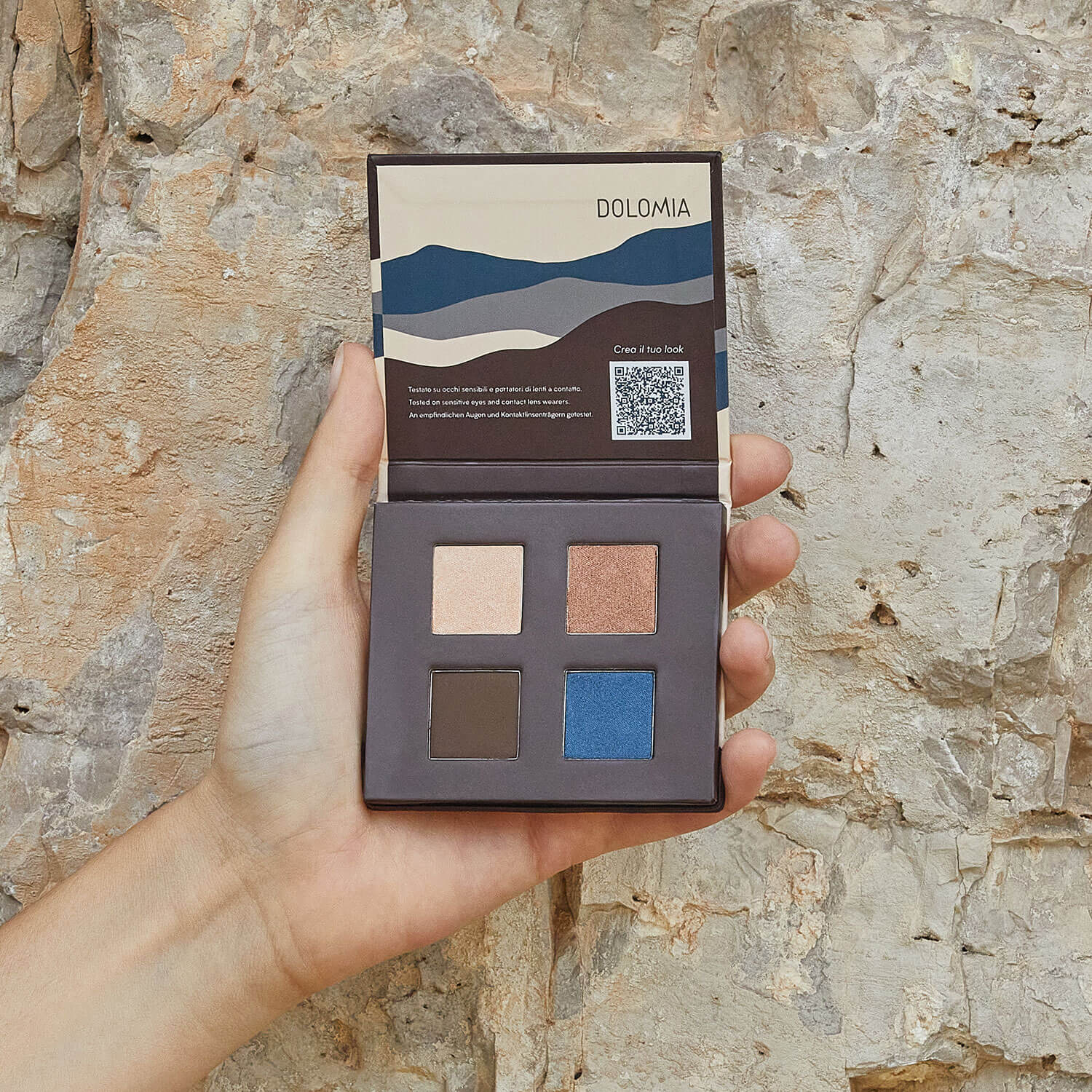 Eine Hand hält eine geöffnete Natural Lidschattenpalette Notte Stellata mit Beige-, Bronze-, Braun- und leuchtenden Dolomitblau-Tönen vor einem strukturierten Steinhintergrund.