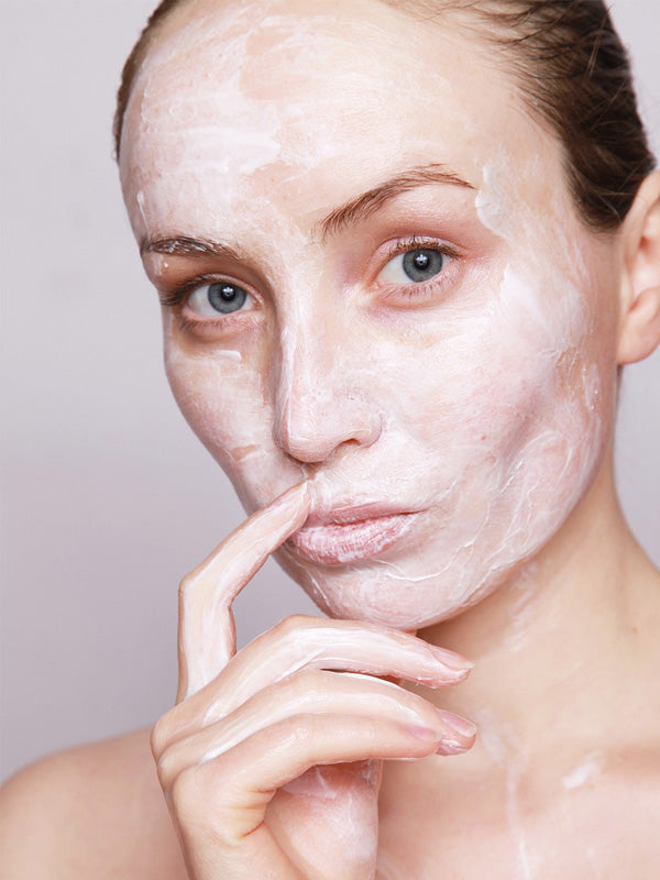 Eine Person mit heller Haut und blauen Augen hat ihr Gesicht mit einer weißen Crememaske bedeckt. Sie schaut in die Kamera und berührt mit einem Finger sanft ihre Lippen. Der Hintergrund ist schlicht und neutral.