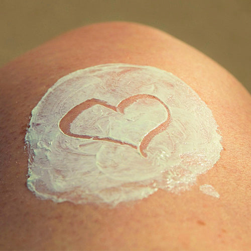 Eine Nahaufnahme der Haut einer Person, auf die weiße Creme aufgetragen wurde. In der Mitte der Creme ist eine Herzform gezeichnet.
