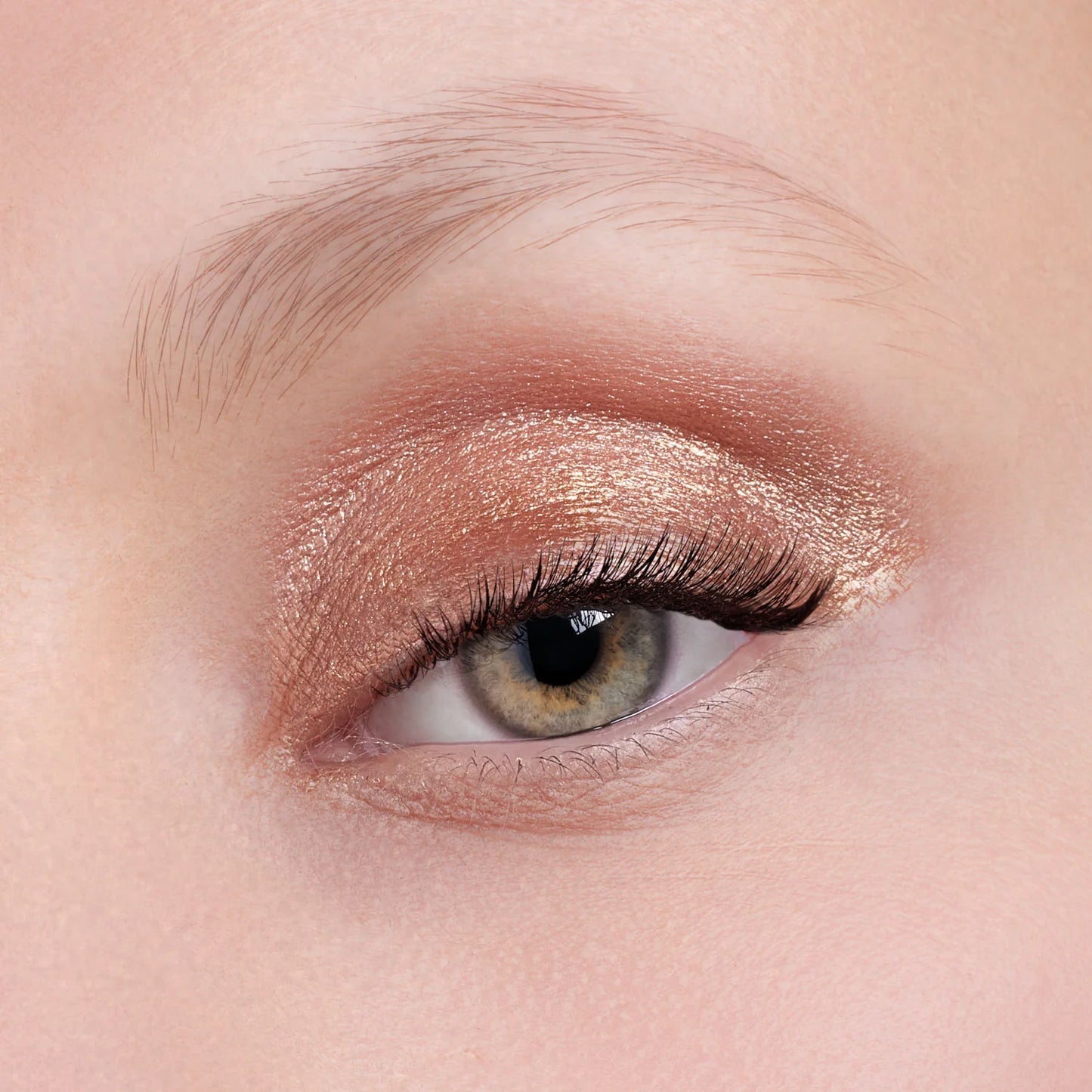 Nahaufnahme eines Auges mit hellgrüner Iris, mit Flüssiger Lidschatten (3 Farbvarianten) in schimmerndem Rosegold-Finish und schwarzer Wimperntusche, darüber eine ausgeprägte Augenbraue.