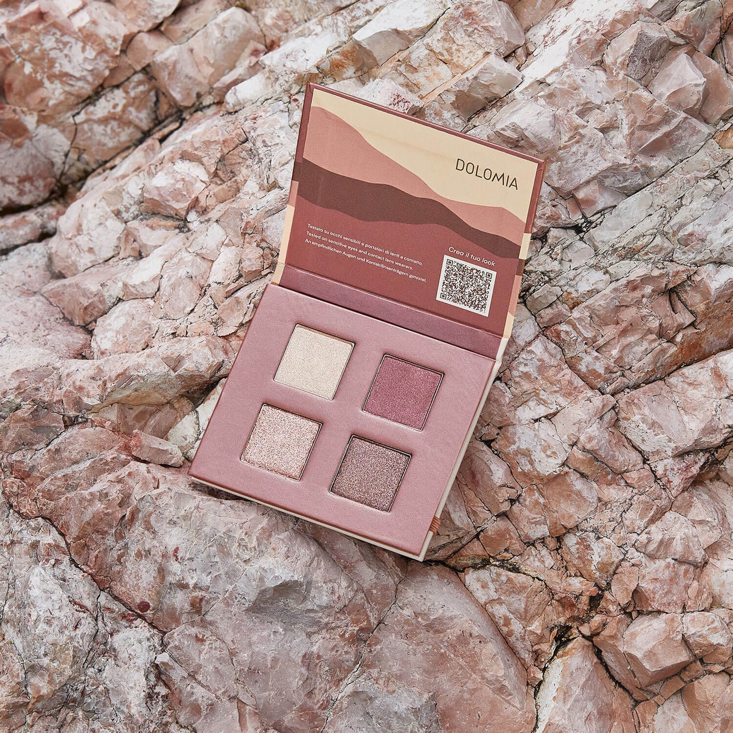 Eine offene Natural Lidschattenpalette Dolomite mit vier glitzernden Rosa- und Beigetönen ruht auf strukturierten hellrosa Felsen. Die Palette zeigt abstrakte Motive und den Markennamen DOLOMIA.