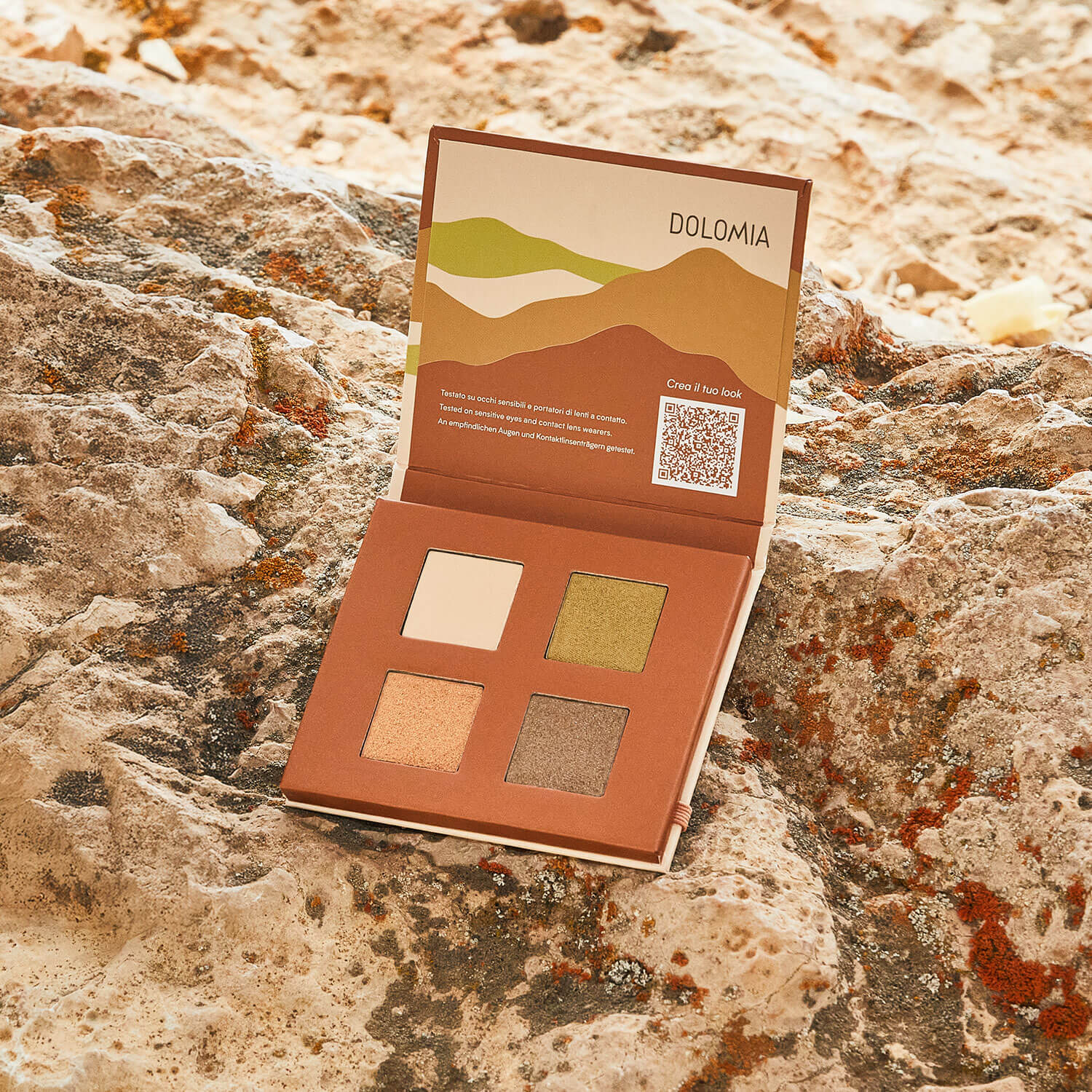 Eine offene Dolomia Natural Lidschattenpalette Botanical mit vier erdigen Farbtönen, darunter Grün- und Goldtöne, ruht auf einer felsigen Oberfläche. Die Innenseite des Covers zeigt abstrakte Landschaftsbilder, Text und einen QR-Code für dauerhafte Tipps.