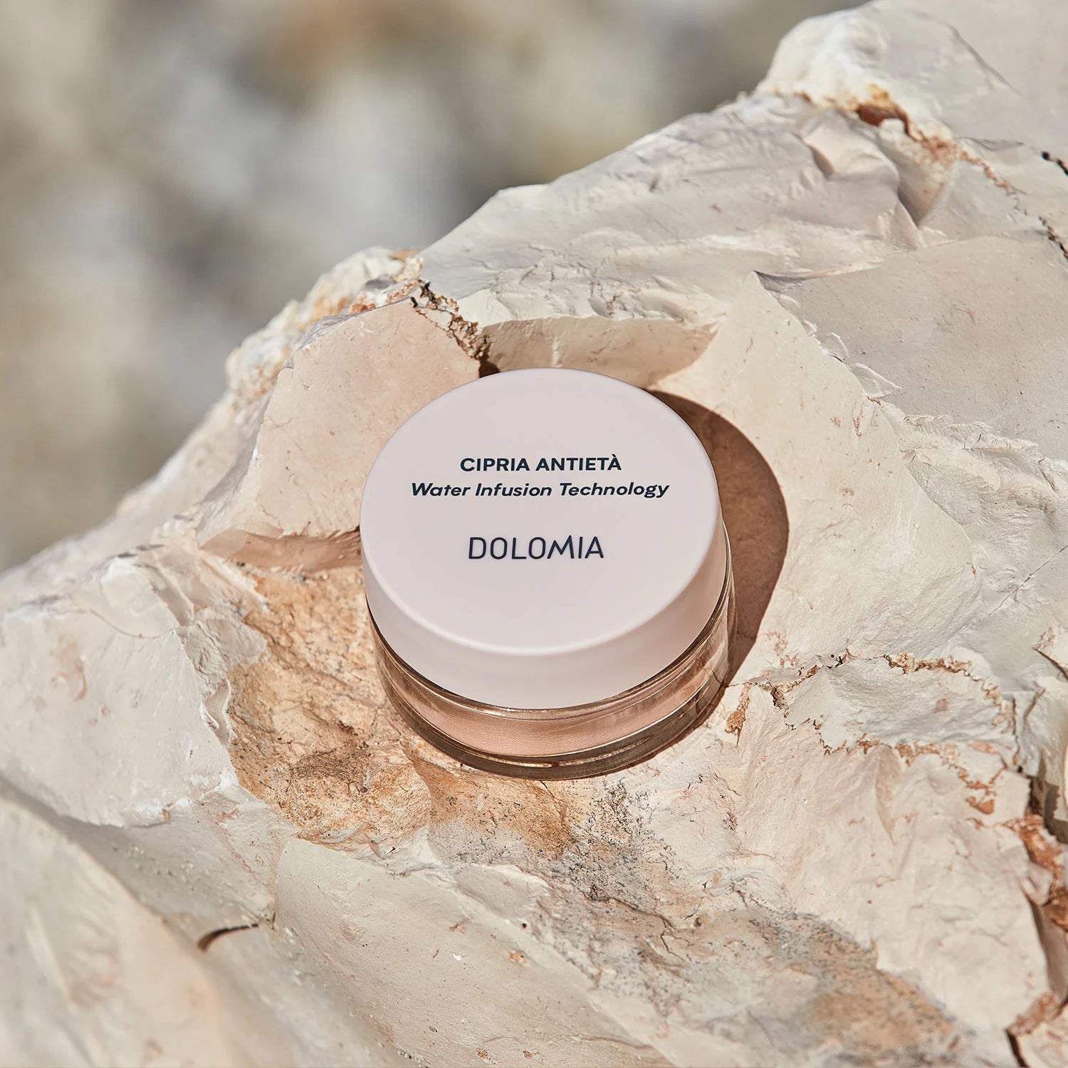 Ein kleiner Tiegel mit Anti-Aging Puder ruht auf einem strukturierten, hellen Stein im natürlichen Sonnenlicht. Auf dem weißen Deckel steht in schwarzer Schrift die Water Infusion Technology für einen makellosen Teint.
