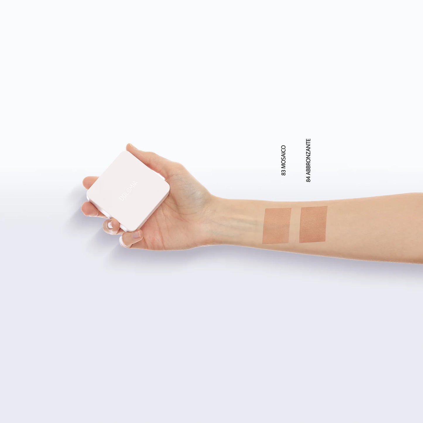 Eine Hand hält die Natural Glow Bronzer-Dose mit zwei braunen Mustern - Moskau und Moscownite - auf dem inneren Unterarm. Hergestellt mit natürlichen Inhaltsstoffen für einen strahlenden Teint. Der Hintergrund ist einfarbig weiß.