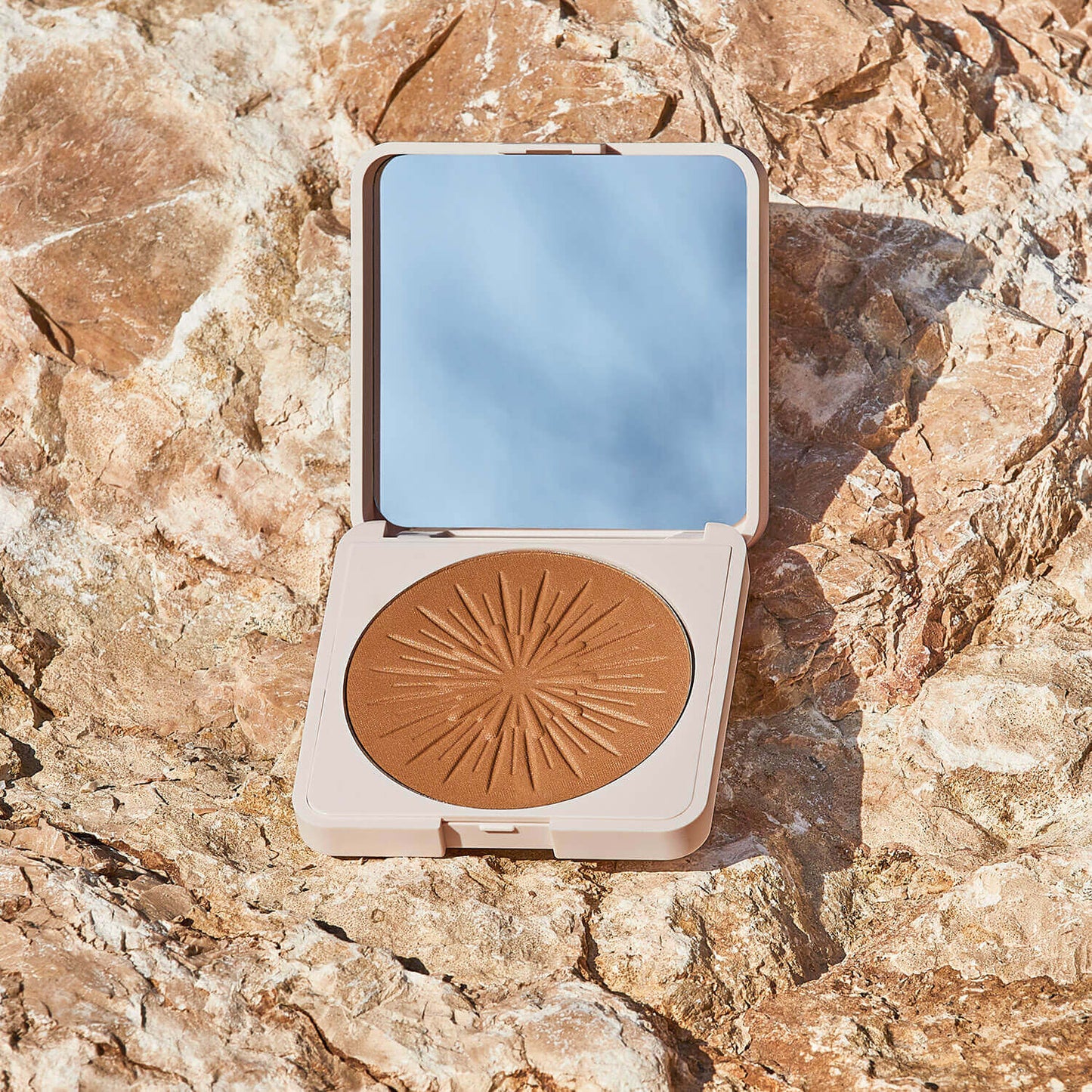 Das Bräunungspuder enthält einen Spiegel und einen gepressten Puder im Sunburst-Design, ideal für einen sonnengeküssten Teint für unterwegs. Abgebildet auf rauen, sonnenbeschienenen Felsen in Hellbraun und Beige - perfekt, um überall einen sonnengeküsster Teint zu erzielen.