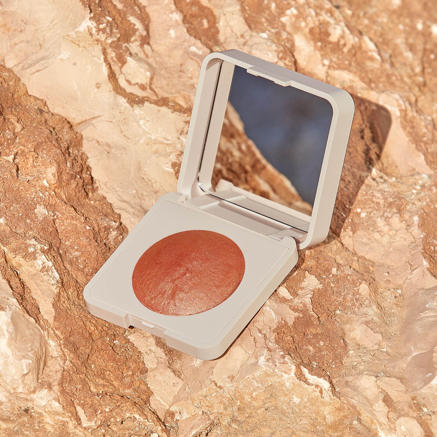 Das Mineral Rouge (2 Farbvarianten), ein kompaktes Make-up mit gebackenem Rouge in einem runden, rötlich-braunen Farbton und eingebautem Spiegel, ruht offen auf strukturiertem Stein - ideal für einen natürlichen Teint.