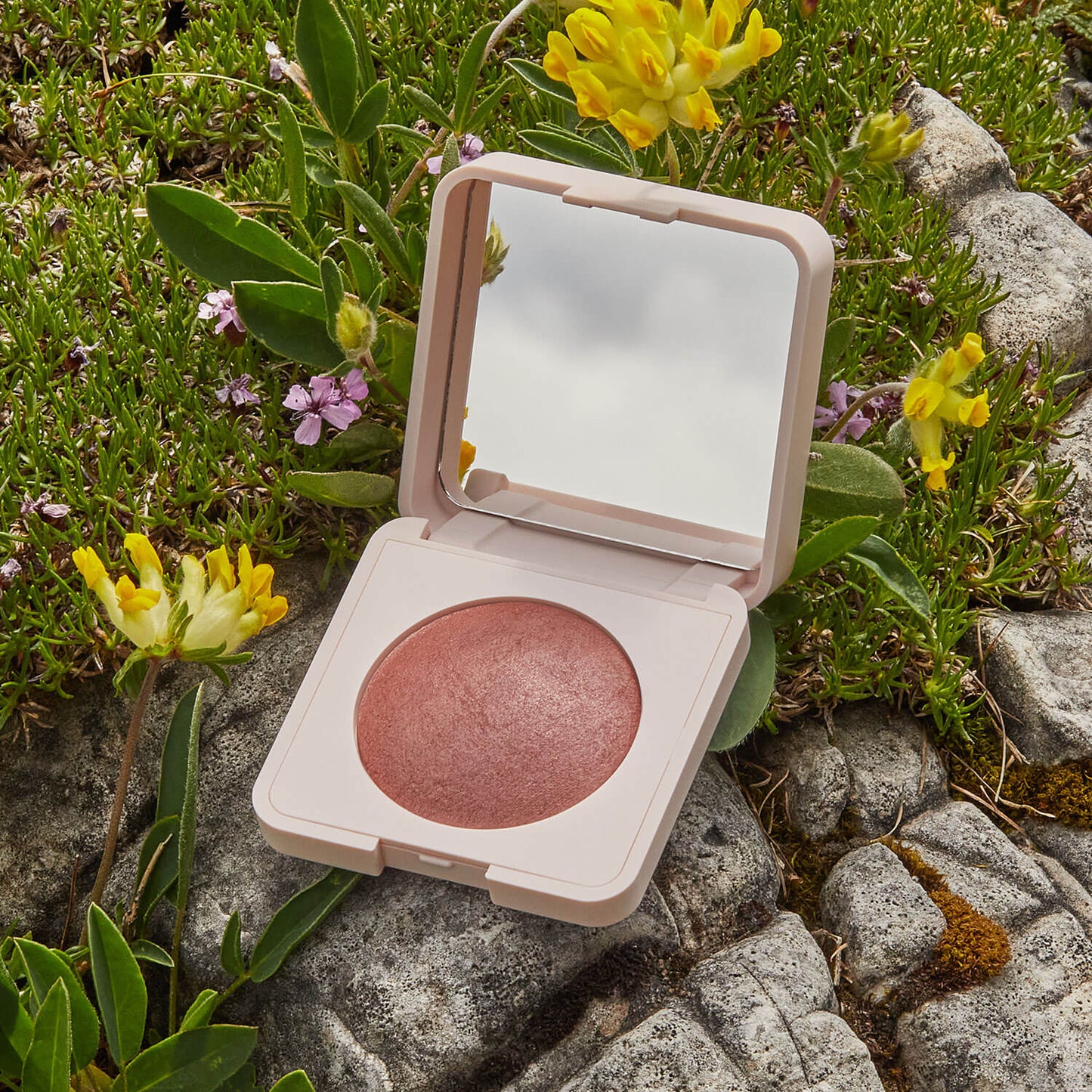 Das Mineral Rouge (2 Farbvarianten) mit Spiegeldeckel liegt offen auf felsigem Grund zwischen grünen Pflanzen und gelben Blüten und sorgt mit seinem schimmernden Rosaton für einen natürlich strahlenden Teint.