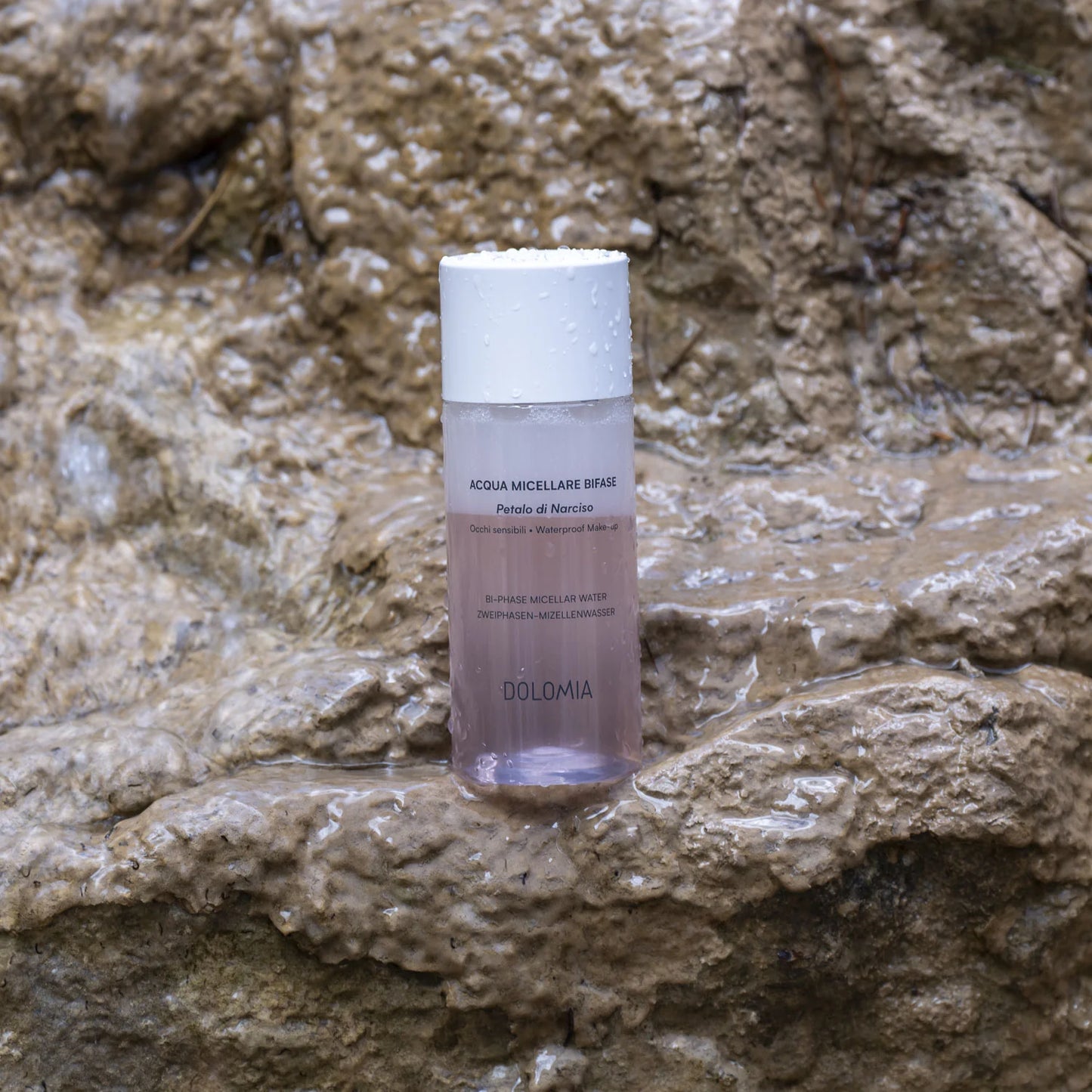 Eine Flasche Zweiphasen-Mizellenwasser Narzissenextrakt sitzt auf nassen, strukturierten Felsen mit Wassertropfen und schafft eine frische, natürliche Szene, die ideal für empfindliche Haut ist.