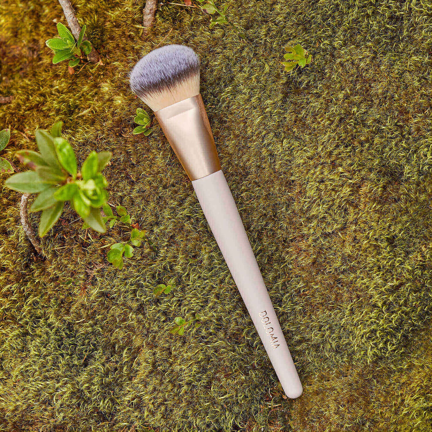 Ein Pinsel für Grundierungen mit goldenem Zwingen und weißem Griff liegt auf grünem Moos, umgeben von kleinen Pflanzen.