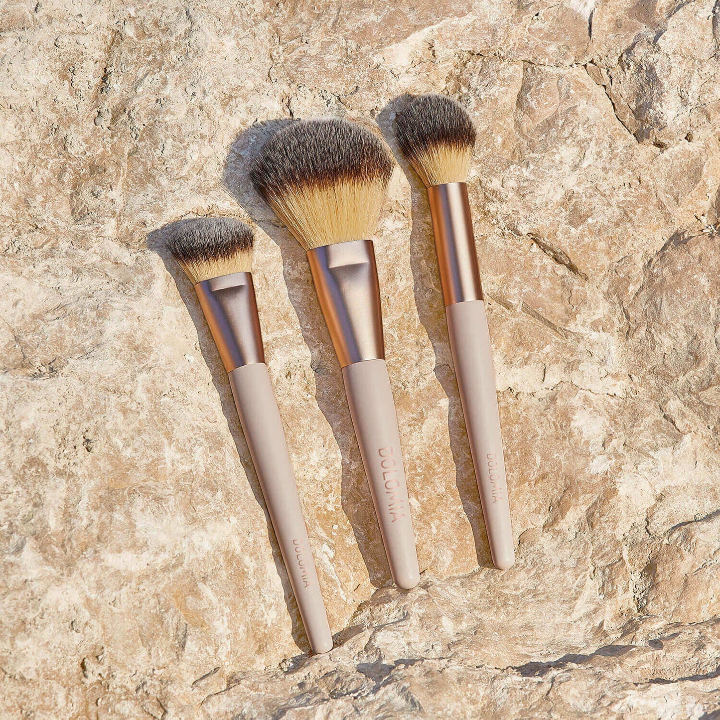 Drei Schminkpinsel, darunter der Rougepinsel mit beigem Stiel und Goldzwinge, sind auf einem strukturierten Stein im Sonnenlicht angeordnet. Alle verfügen über hochwertige synthetische Pinselhaare für eine präzise Gesichtskontur.