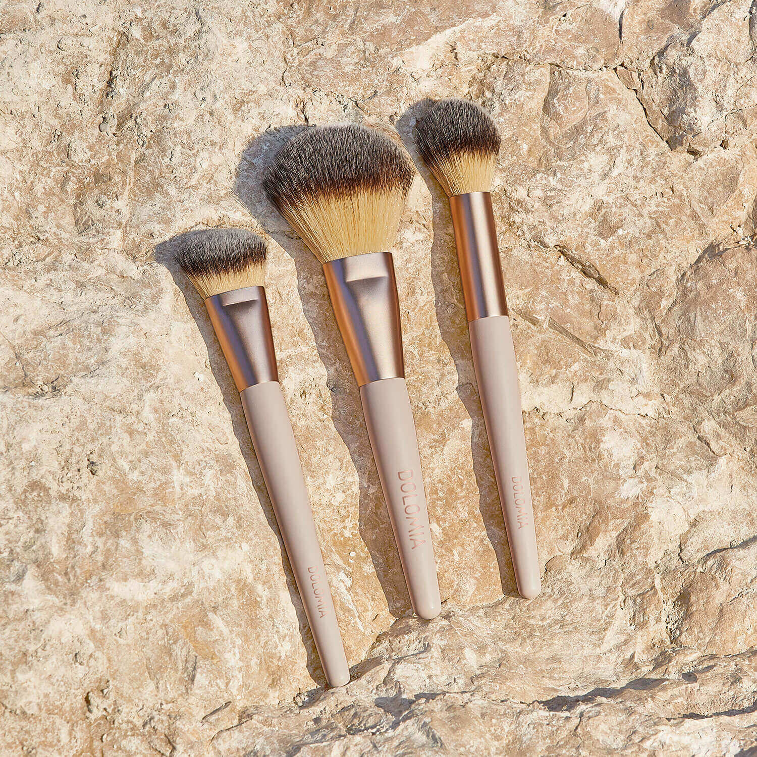 Drei Schminkpinsel, darunter der Rougepinsel mit beigem Stiel und Goldzwinge, sind auf einem strukturierten Stein im Sonnenlicht angeordnet. Alle verfügen über hochwertige synthetische Pinselhaare für eine präzise Gesichtskontur.