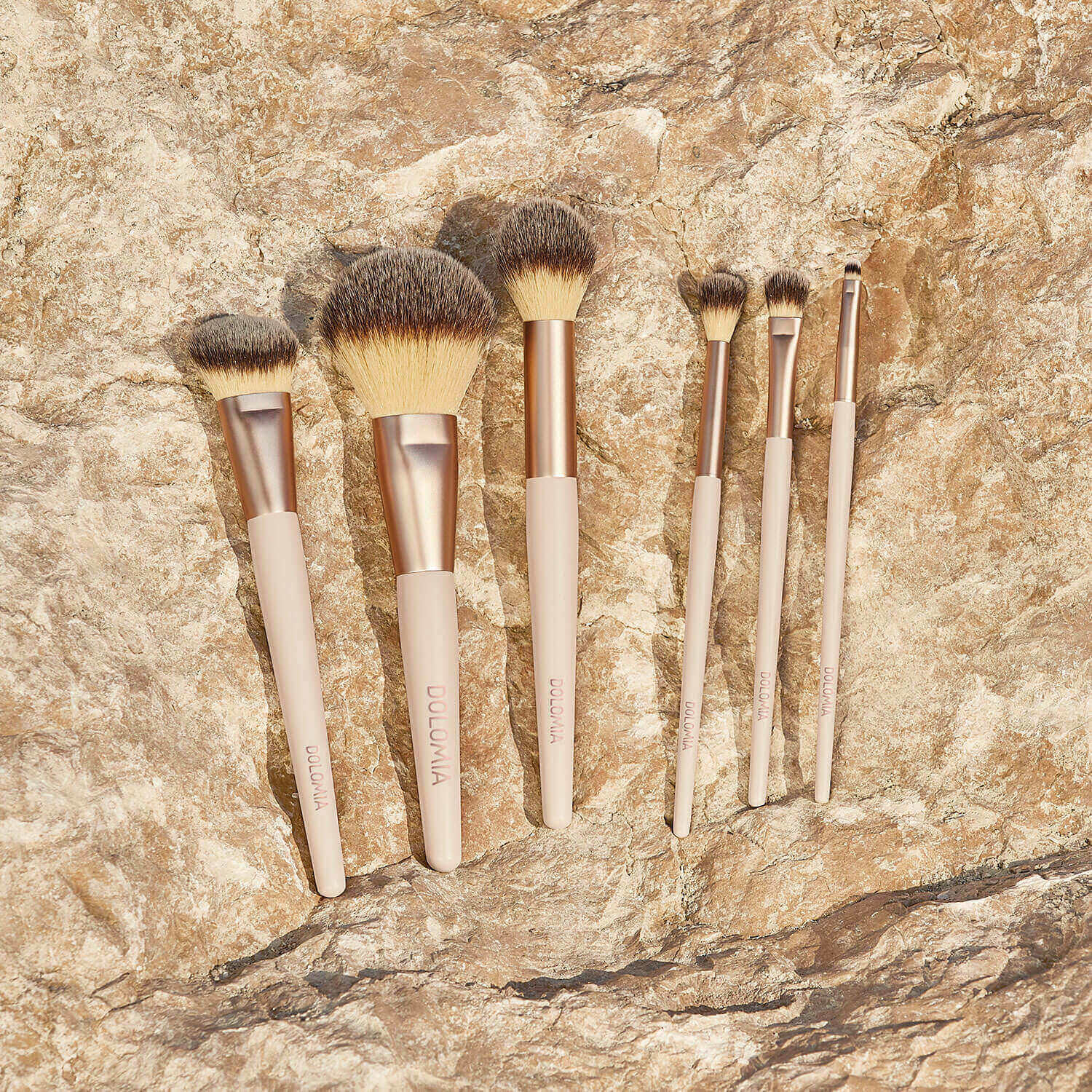 Sechs Augenpinsel für Schattierungen mit beigen Griffen und goldenen Zwingen, darunter Lidschattenpinsel, sind in einer Reihe auf einer sonnenbeschienenen Steinfläche angeordnet. Alle verfügen über synthetische Fasern für einen reibungslosen Make-up-Auftrag.