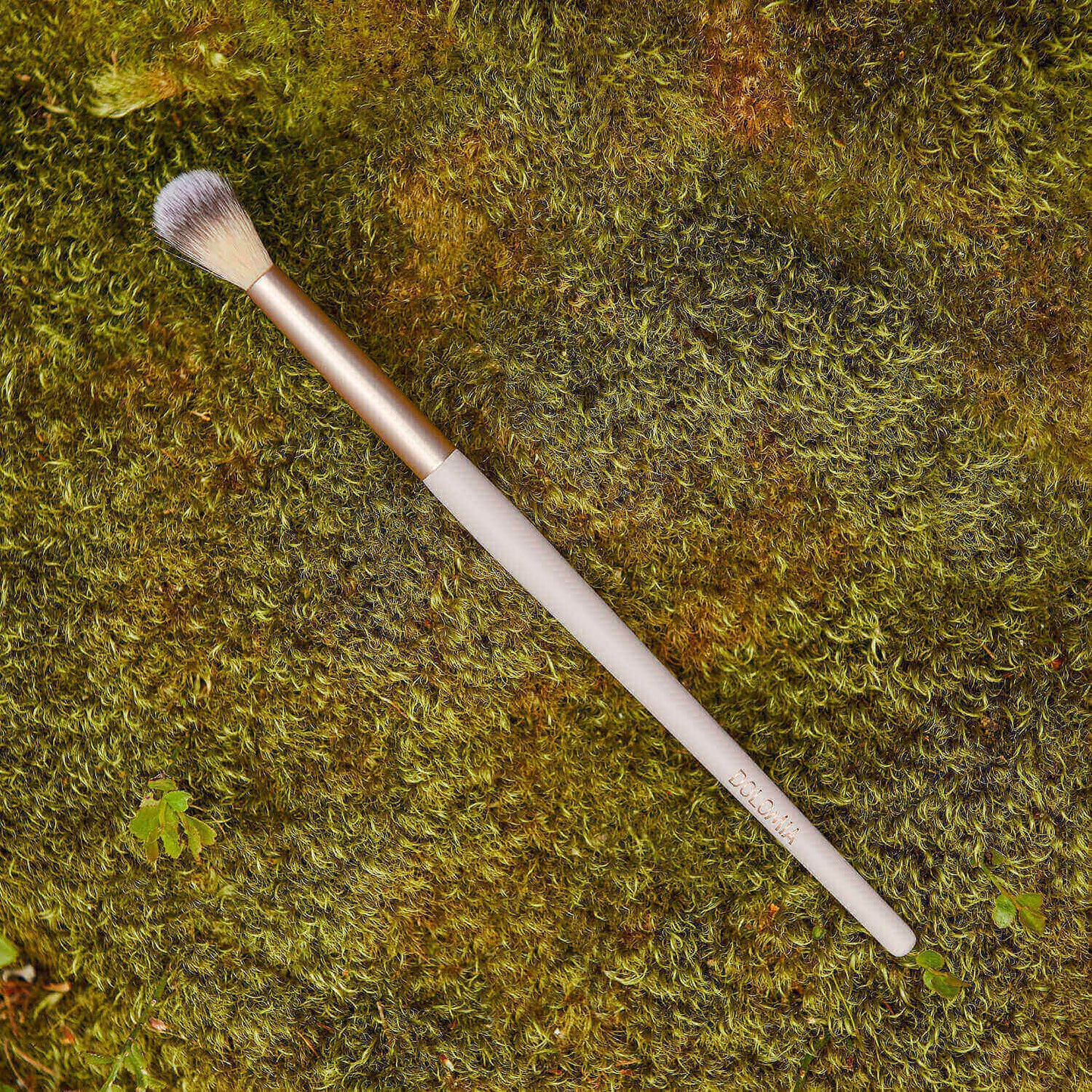 Der Augenpinsel für Lichteffekte mit leichtem Griff und flauschigen Synthetikborsten liegt schräg auf grünem Moos - ideal zum Verblenden von Lidschatten oder präzisem Augen-Make-up.