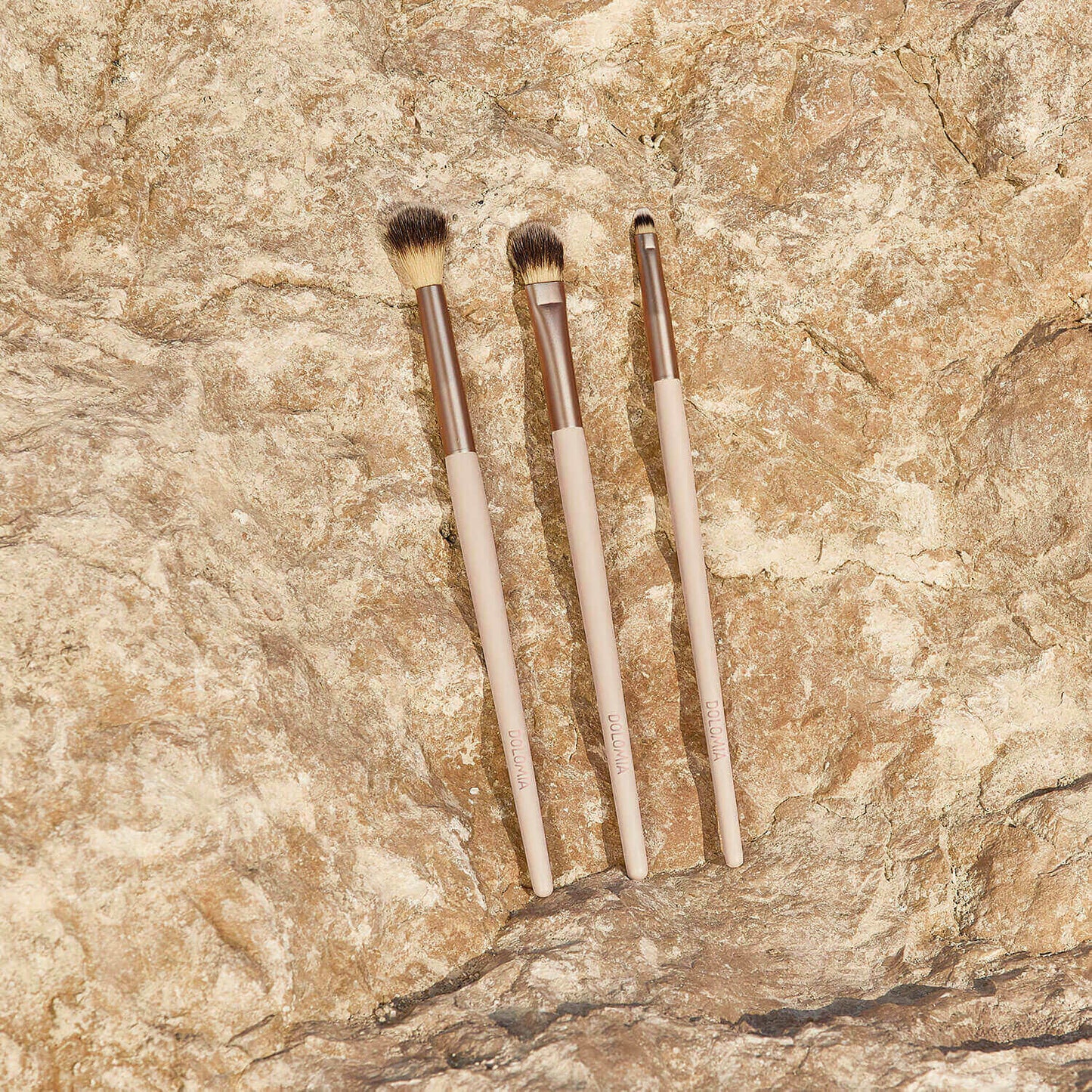 Drei Augenpinsel für Schattierungen mit beigen Griffen und braunen Metallzwingen, hergestellt aus synthetischen Fasern, sind nebeneinander auf einer strukturierten, hellbraunen Steinoberfläche platziert.