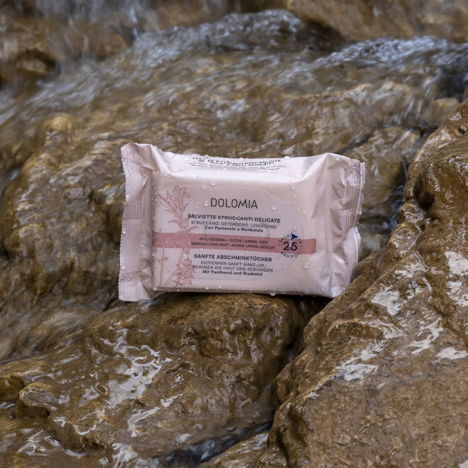 Eine Packung Sanfte Abschminktücher Augen-Lippen-Gesicht für empfindliche Haut liegt auf nassen Steinen, in deren Nähe Wasser wirbelt. Die hellrosa, blumige Verpackung enthält 25 Abschmink-Tücher.